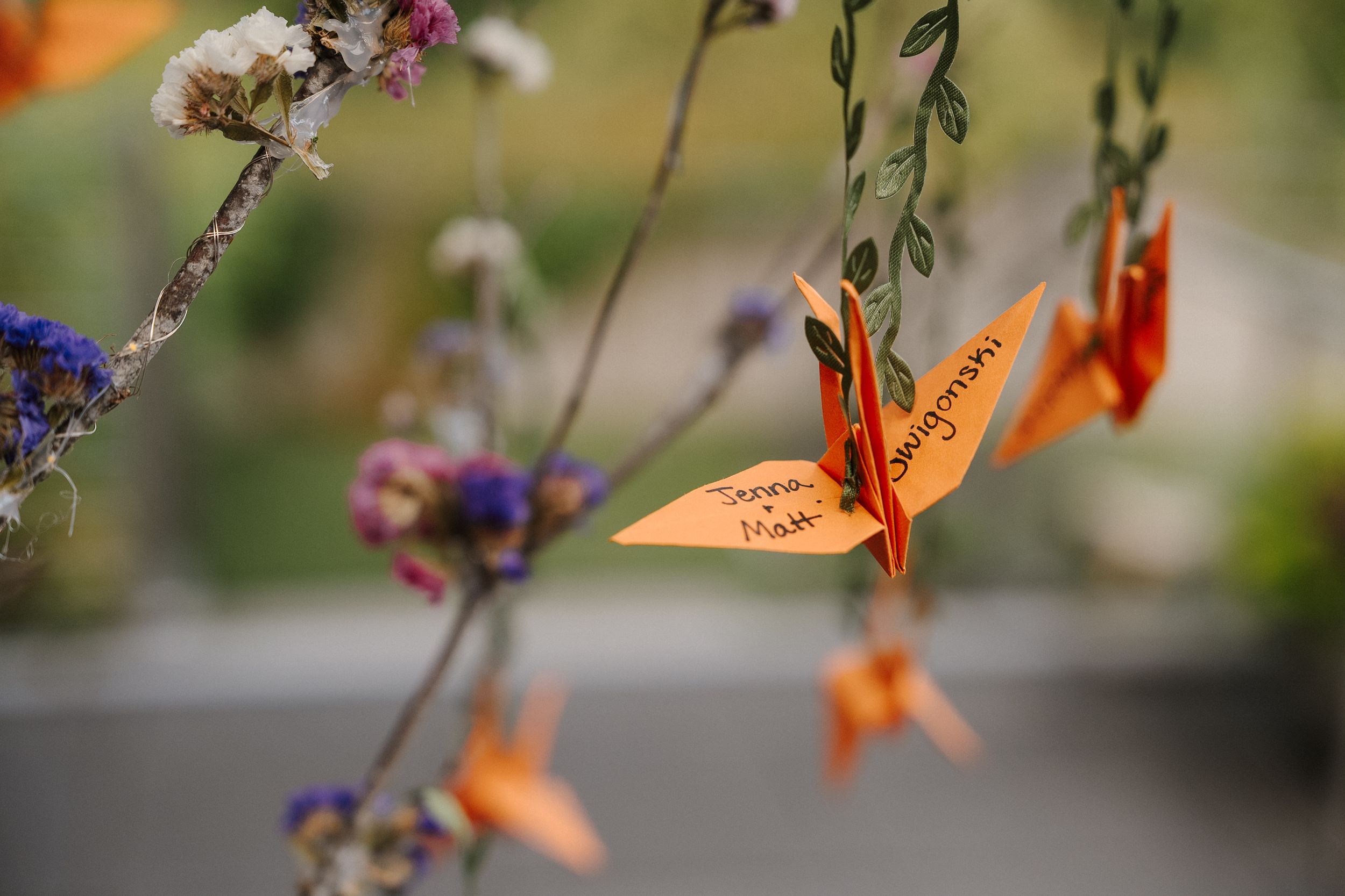 Delicate orange origami paper cranes hanging from flowering tree branches against a blurred natural background.