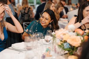 Wedding guests laugh and smile during a reception dinner at a decorated table with floral centerpieces.