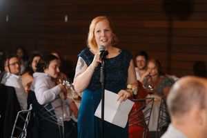 A speaker in a blue dress addresses an audience during an event while holding a microphone and papers.