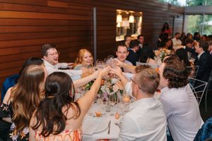 Wedding guests sitting at long tables raise their glasses in a celebratory toast during a reception.