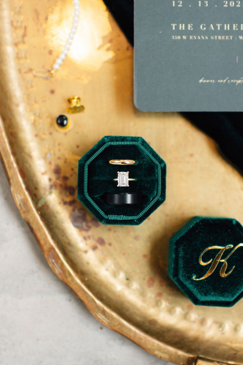 Close-up of emerald green velvet ring boxes and wedding stationery on gold metallic surface.