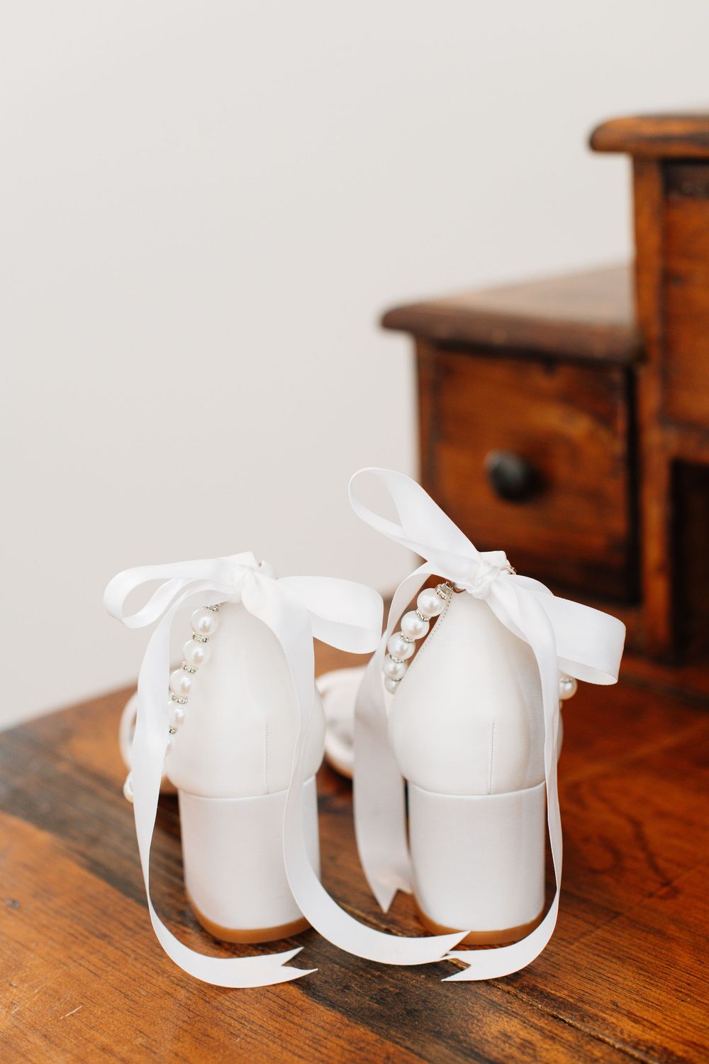 White vintage-style bridal shoes with ribbon bows on wooden furniture.