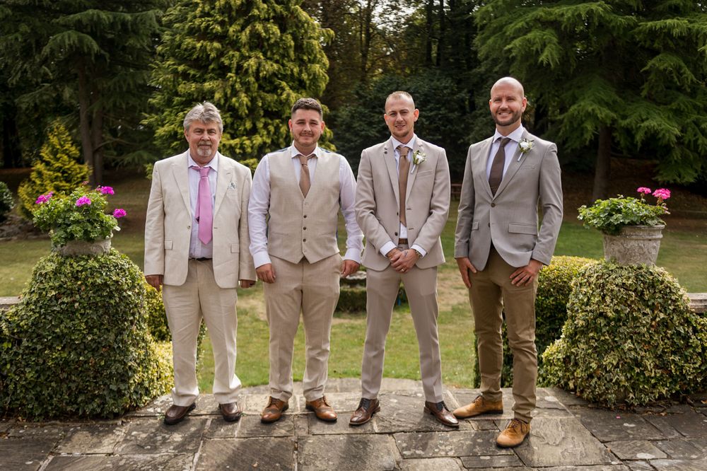 Groomsmen in light colored suits stand together outdoors showing off their dress shoes.