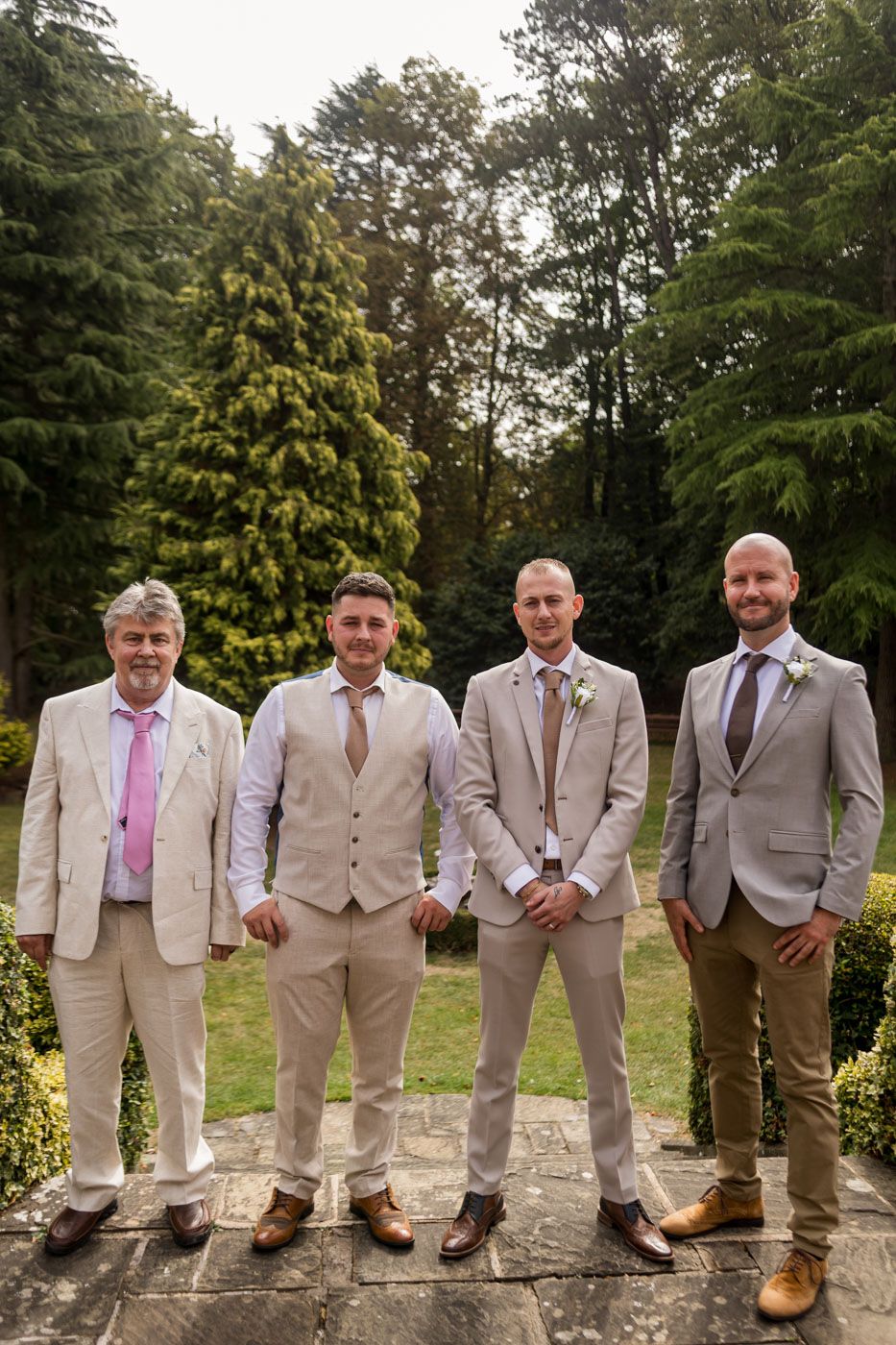 Groomsmen lined up outdoors wearing coordinated beige suits and brown shoes for wedding photos.