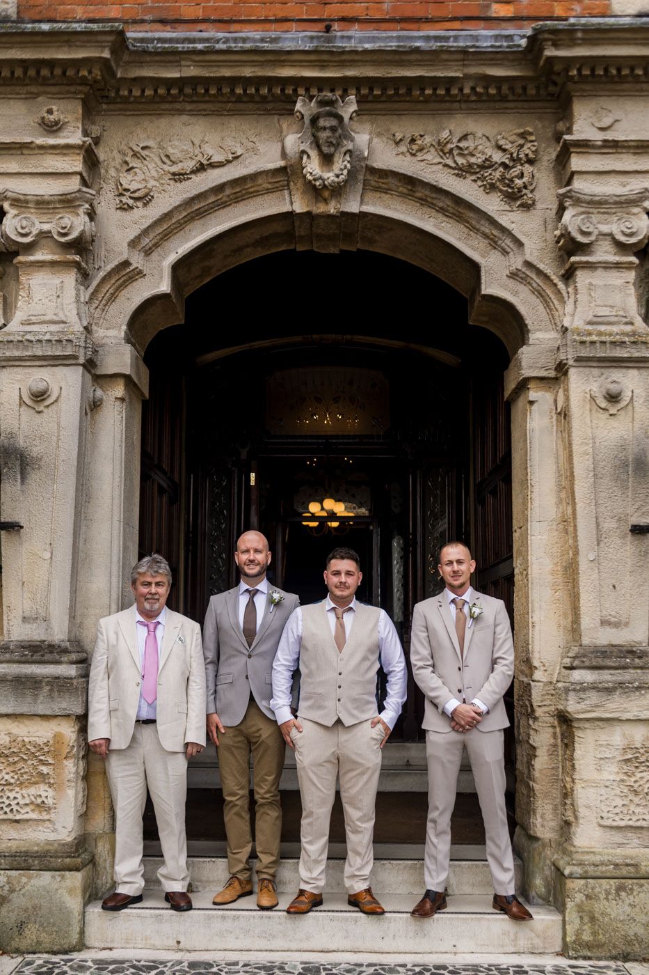 Wedding party poses in formal attire under ornate stone archways of historic building.