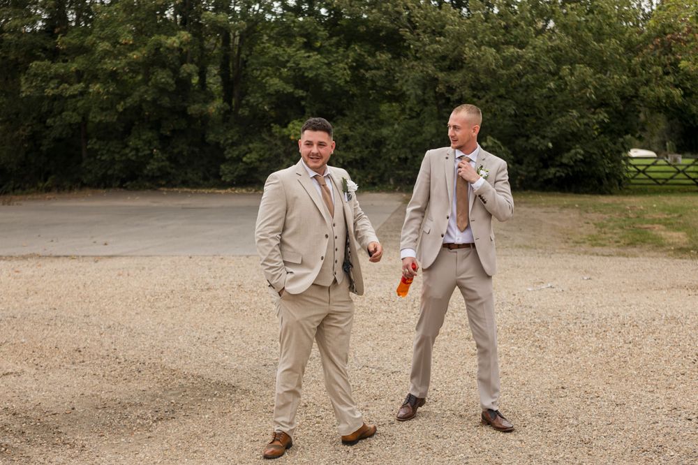 Groomsmen in beige suits walk together on gravel path during wedding photos.