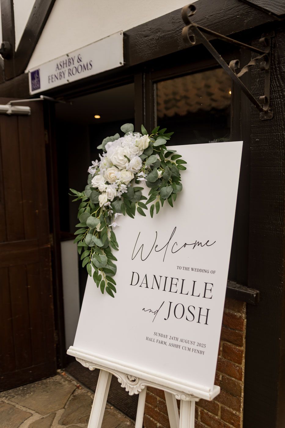 Elegant white wedding welcome signs decorated with greenery displayed at venue entrance.