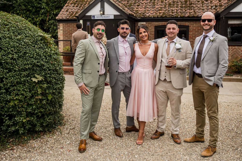 Wedding party in light colored suits and pink dress pose together outside a rustic brick building.
