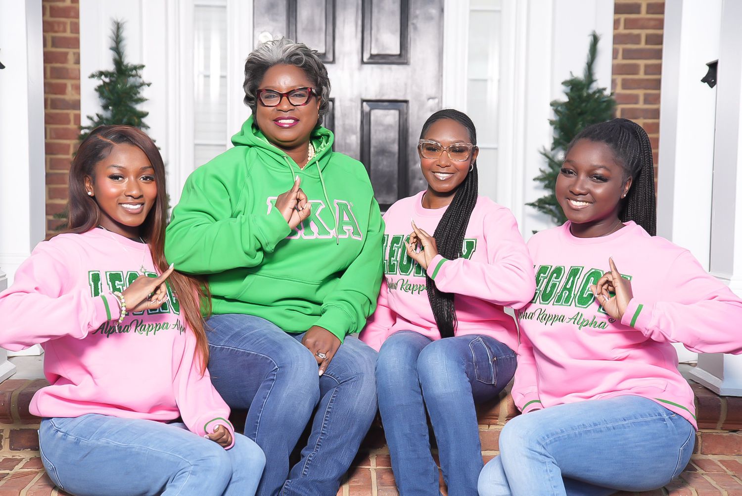 Four women in pink and green sorority sweatshirts pose with hand signs while sitting on porch steps near a front door.