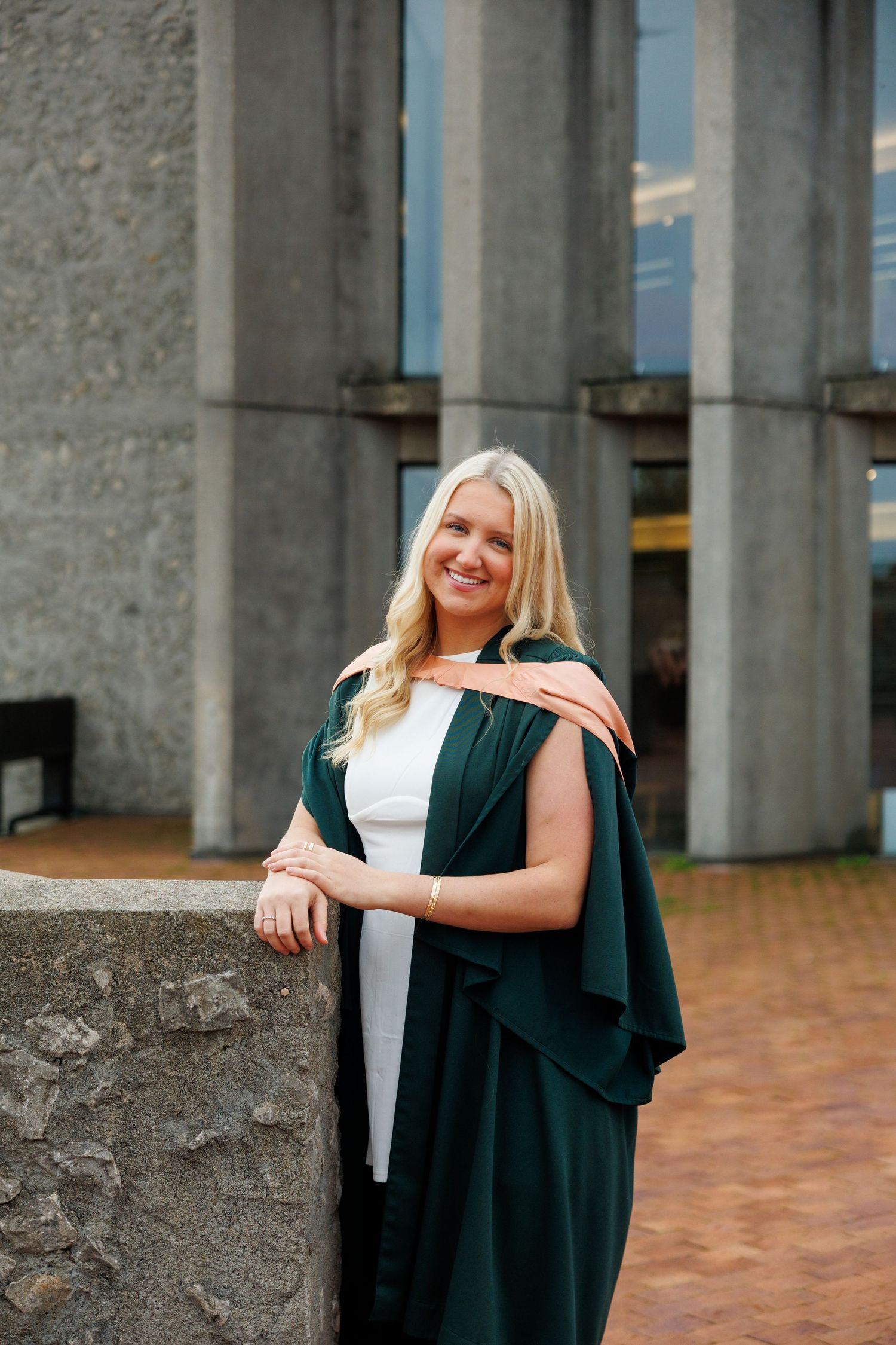 Graduate in black academic regalia poses against stone columns during outdoor graduation photo session.