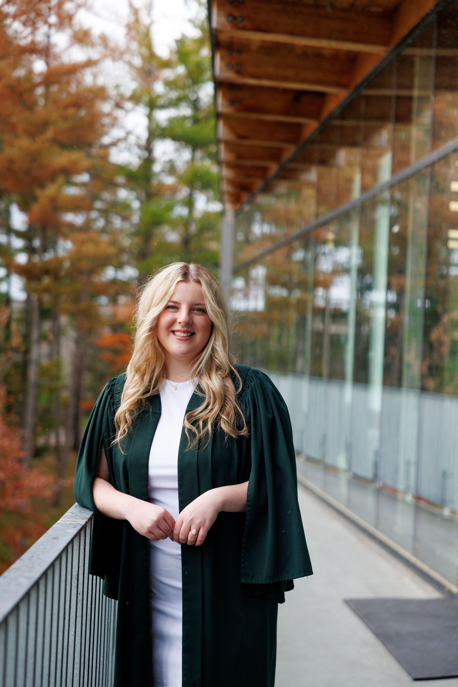 Graduate posing in black academic gown on outdoor walkway with autumn foliage in background.