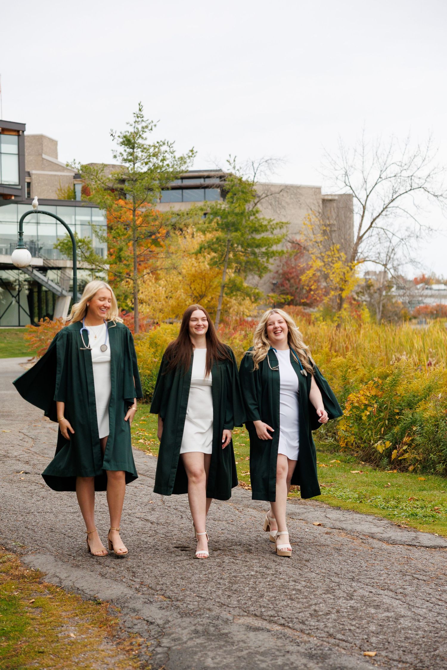 Four friends in matching green blazers and white dresses walk along an autumn path beside modern buildings.