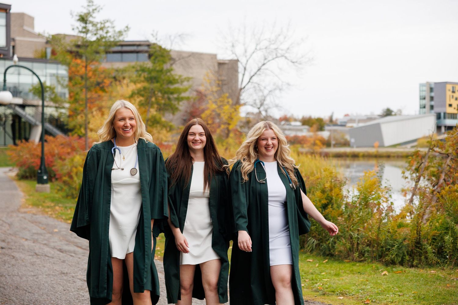 Three people in graduation attire smile and laugh together while walking outdoors on a fall day.
