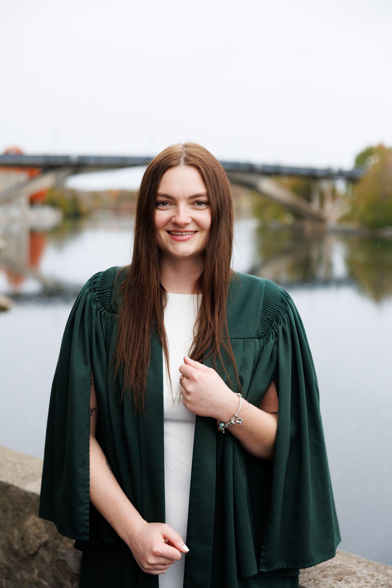 Graduate in emerald green academic regalia poses outdoors near a waterfront on an autumn day.