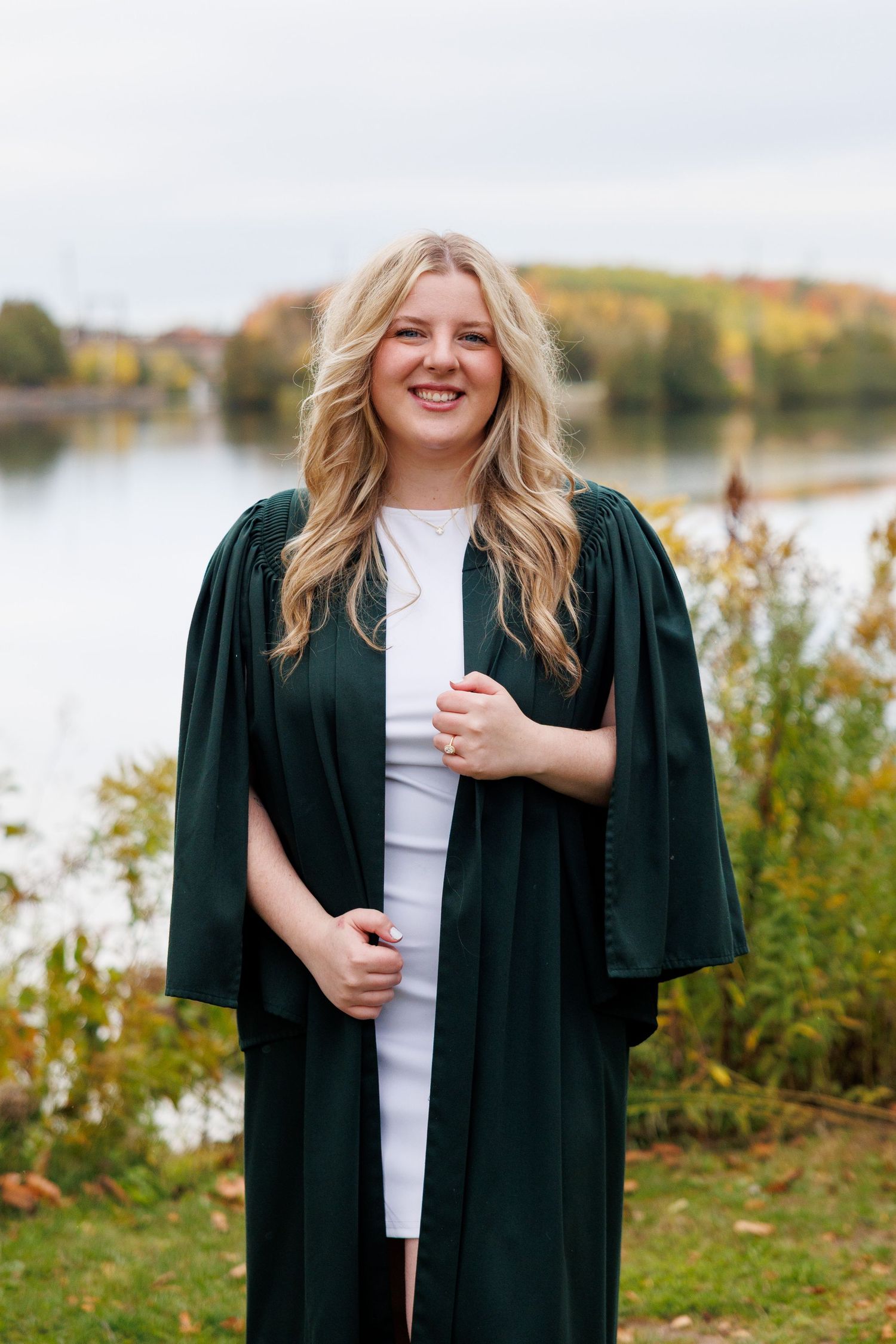 A person in graduation attire poses outdoors near a lake during autumn with fall foliage in the background.