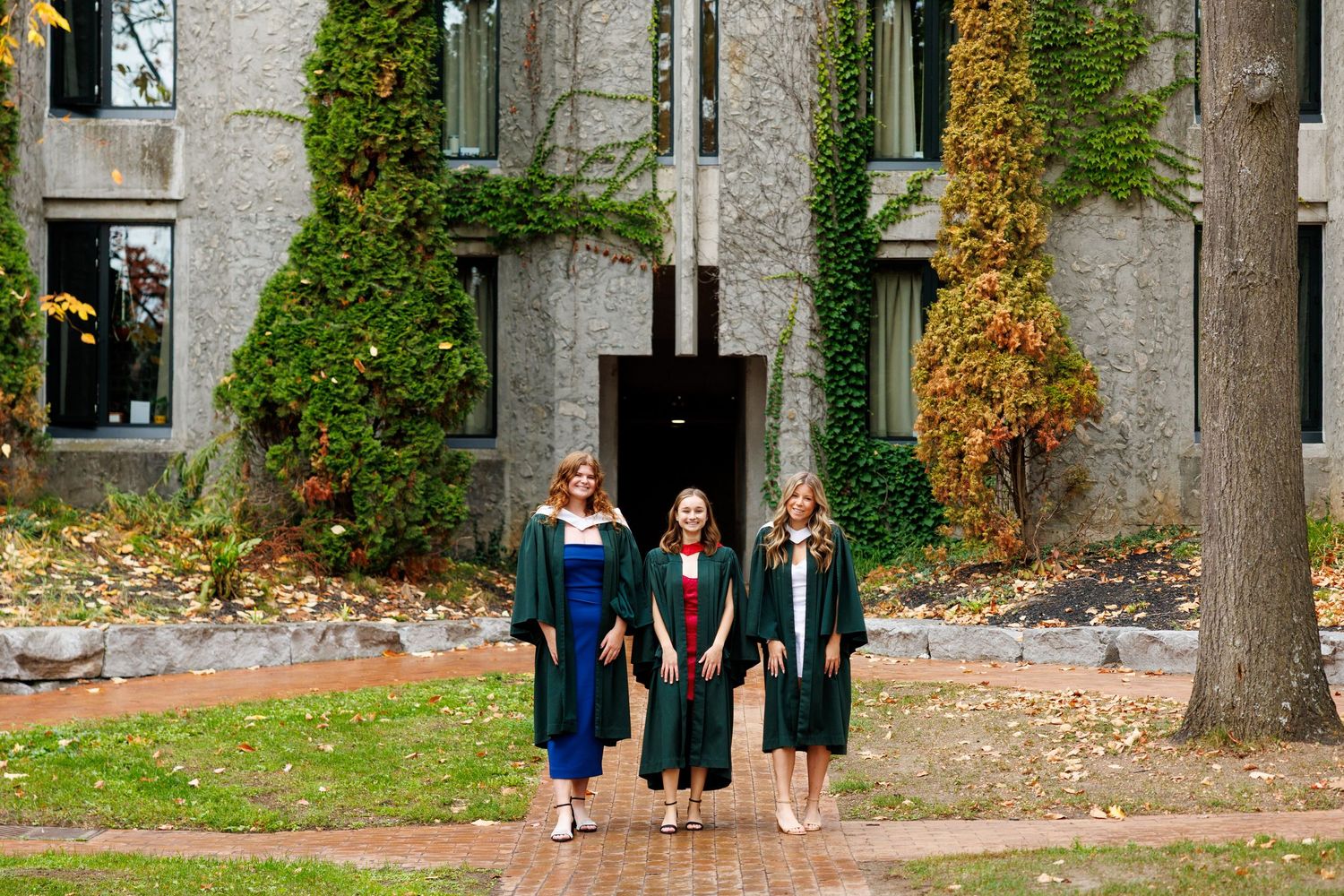 Three people in graduation attire walk near a stone building with autumn foliage.