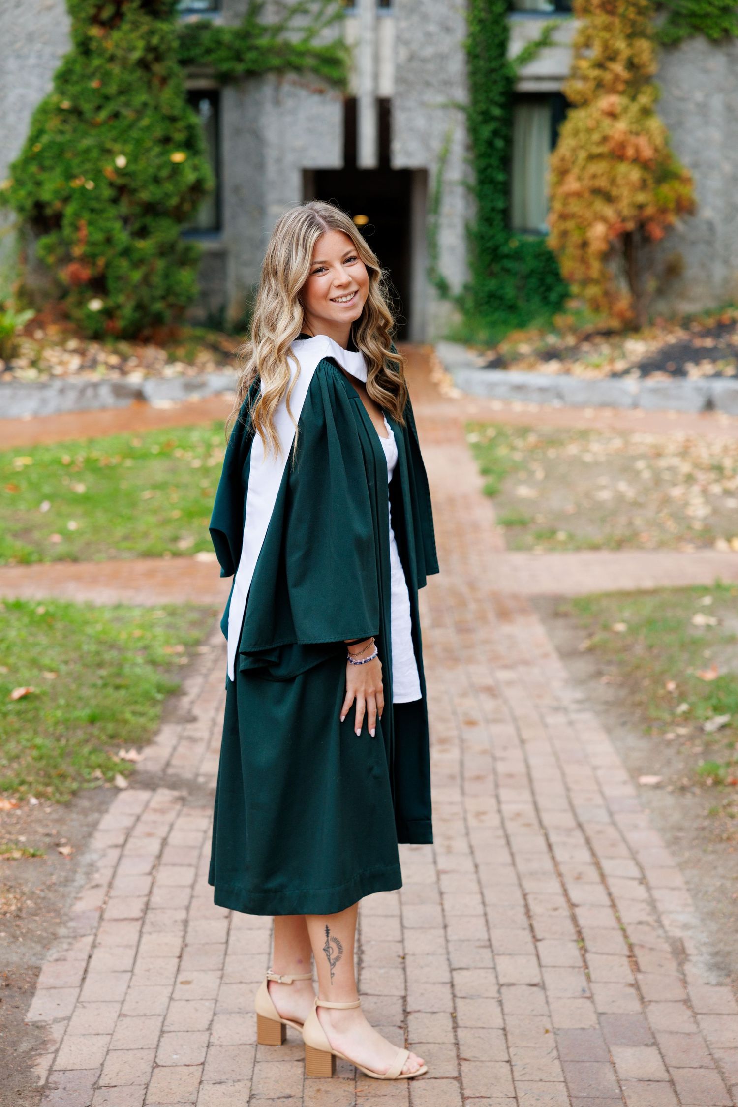 A graduate in a dark green academic gown walks along a brick pathway during a fall graduation photoshoot.