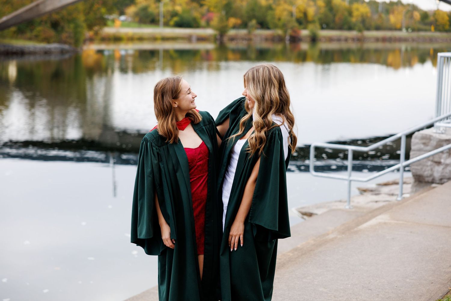 Two people in graduation gowns stand together laughing by a waterfront on a fall day.