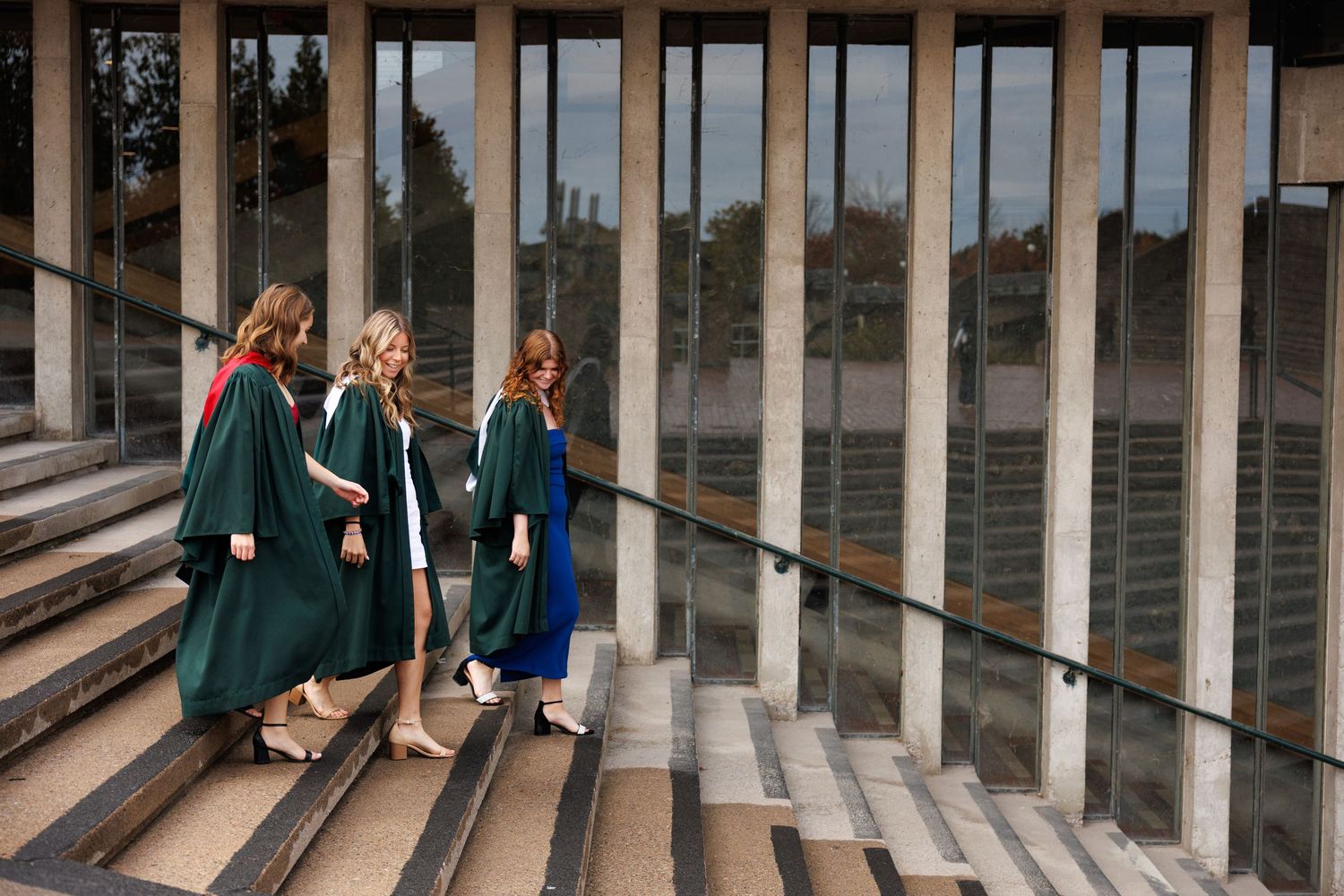 Four graduates in dark robes walk along a stone colonnade at a historic building.