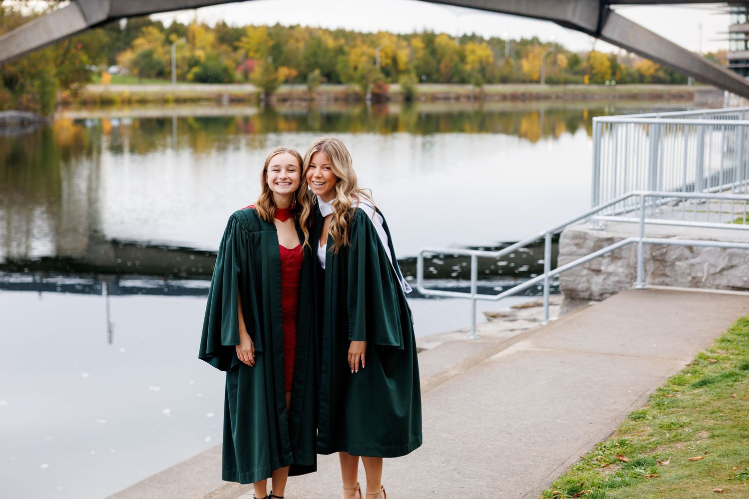 Two graduates in dark green graduation gowns stand together by a lake on a fall day.
