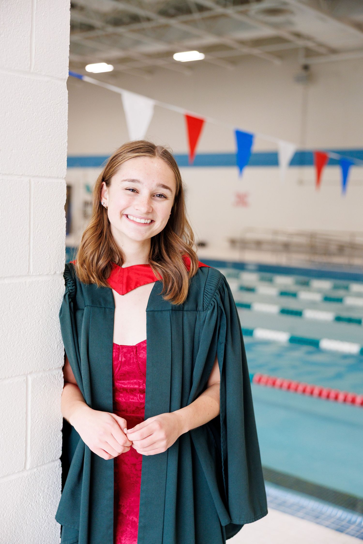 Graduate in green cap and gown smiles while posing by indoor swimming pool with red and blue pennants.