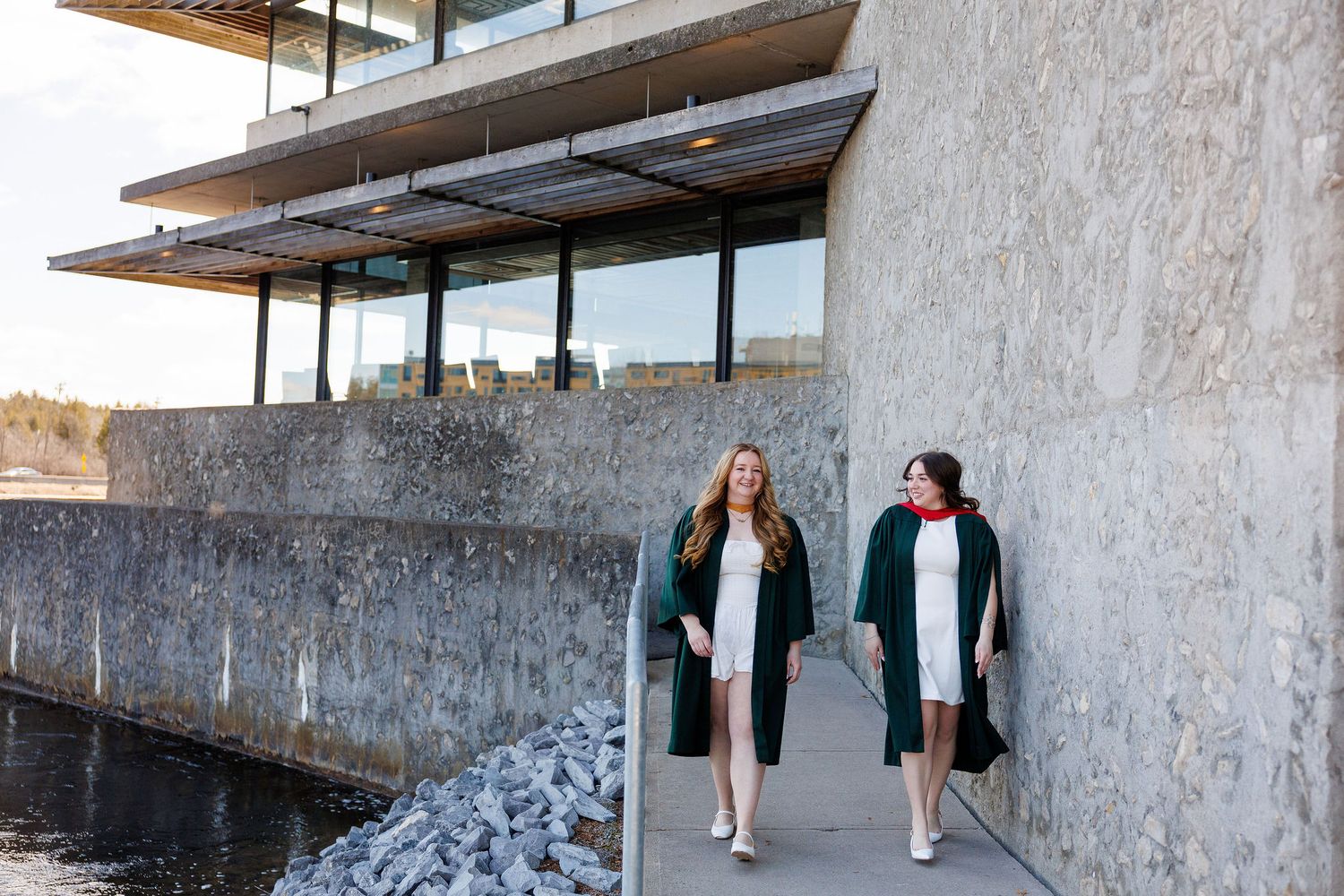 Two graduates in green gowns walk along modern waterfront building exterior.