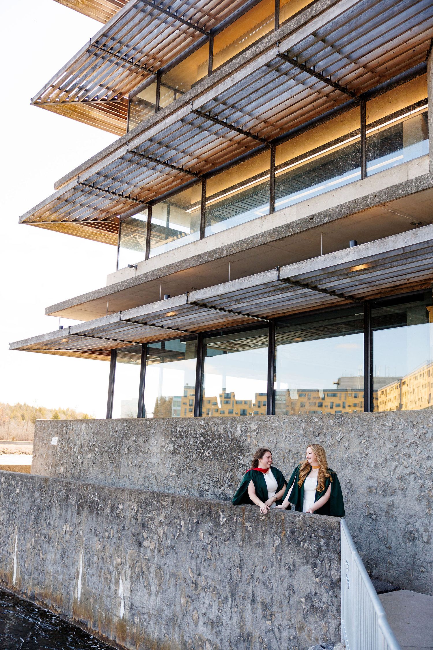 Two graduates in academic gowns stand beside a concrete wall outside a modern multi-story building with wooden canopy details.
