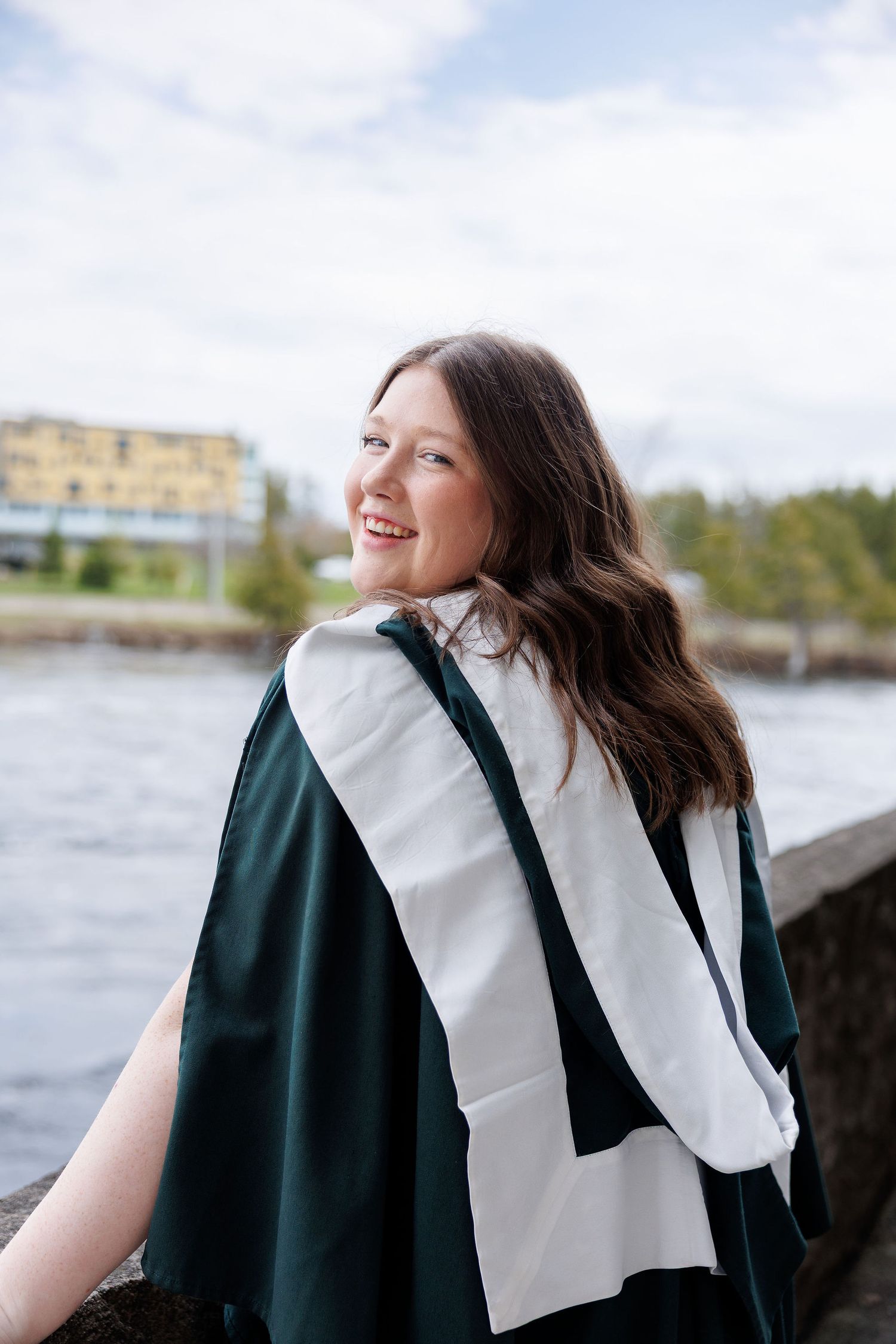 Smiling young woman in green graduation gown posing by a river on a cloudy day.