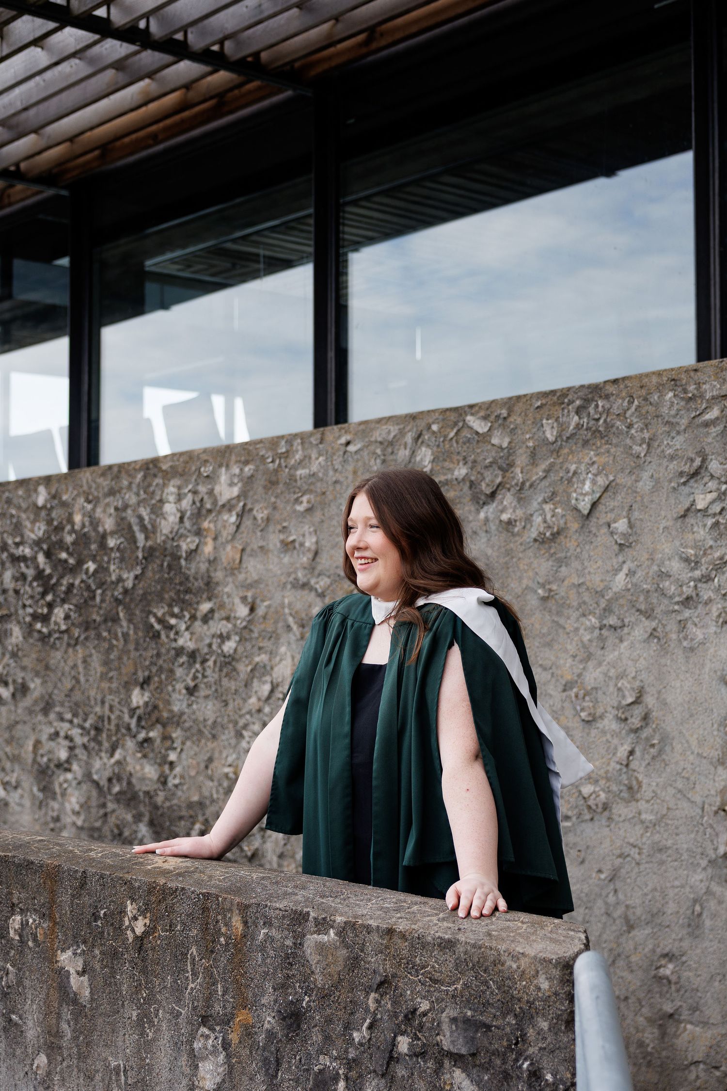 Smiling woman in green graduation gown leaning on stone wall outdoors near modern building windows.