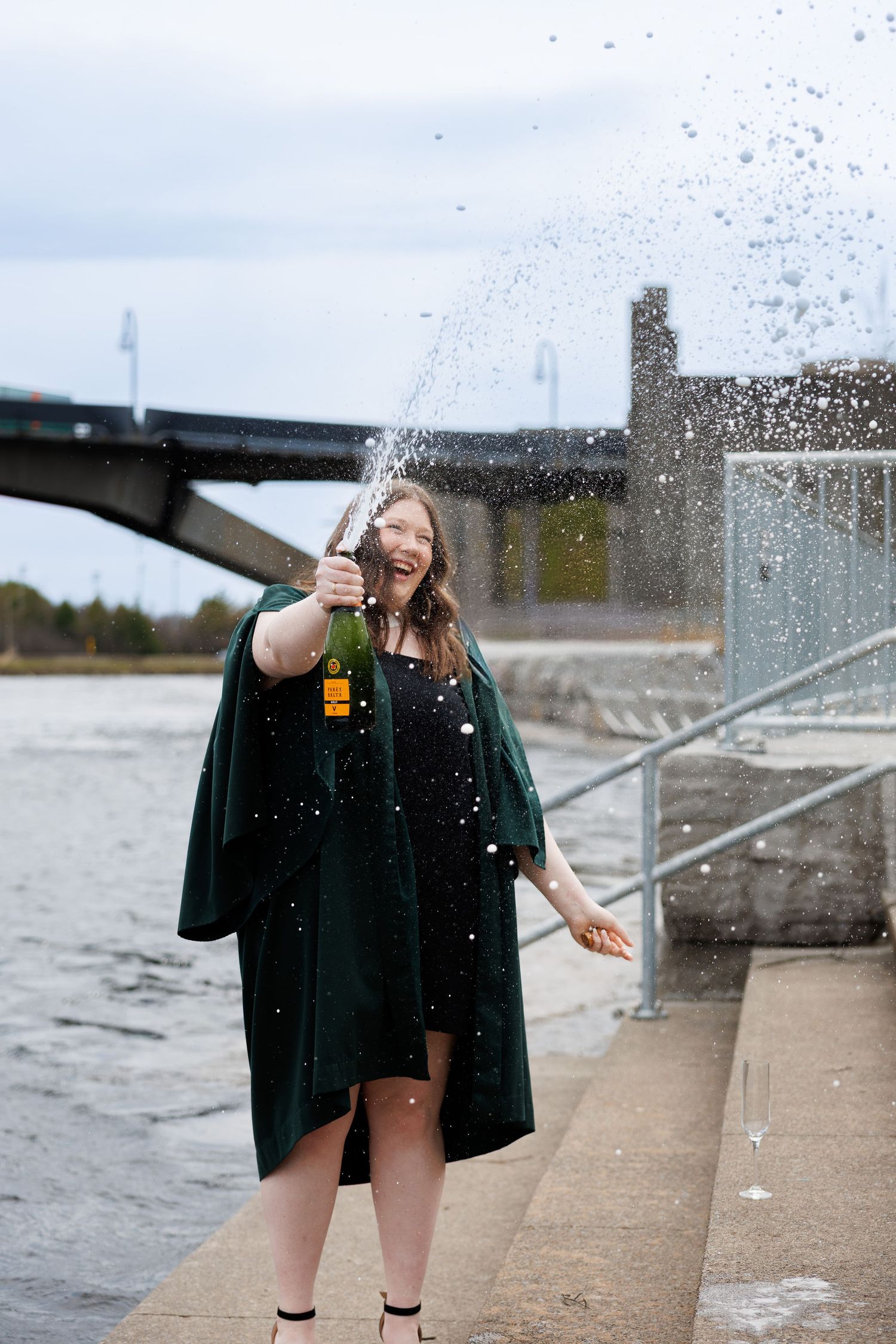 Graduate in green gown celebrates by popping champagne bottle outdoors near waterfront bridge.