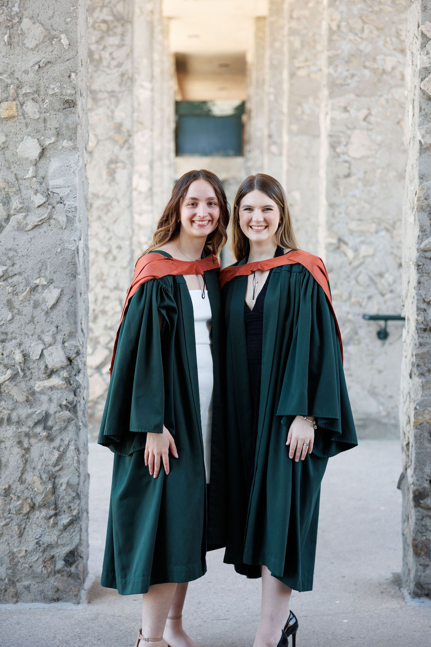 Two smiling women in dark green graduation gowns with red hoods pose together in front of stone columns.