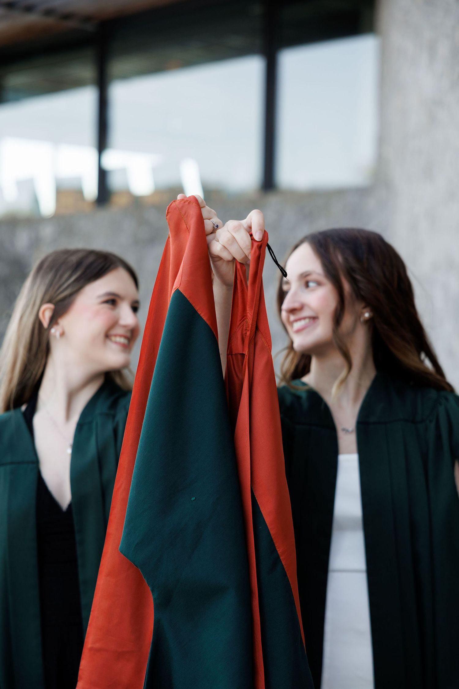 Two smiling graduates in green robes hold red graduation caps outdoors on their graduation day.