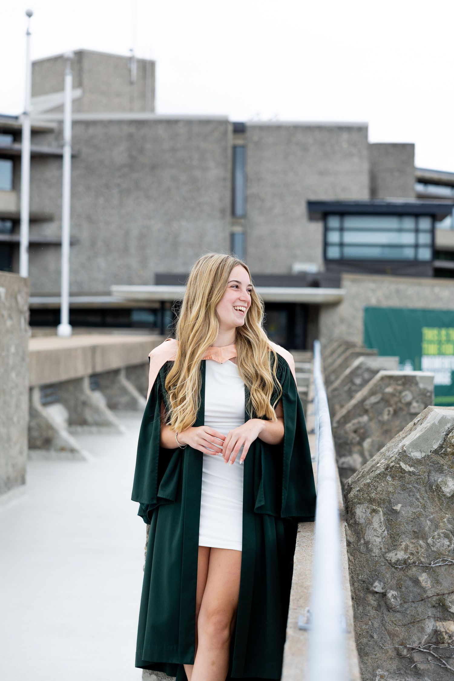 Smiling blonde woman in white dress and dark green graduation gown standing outdoors near a modern building.