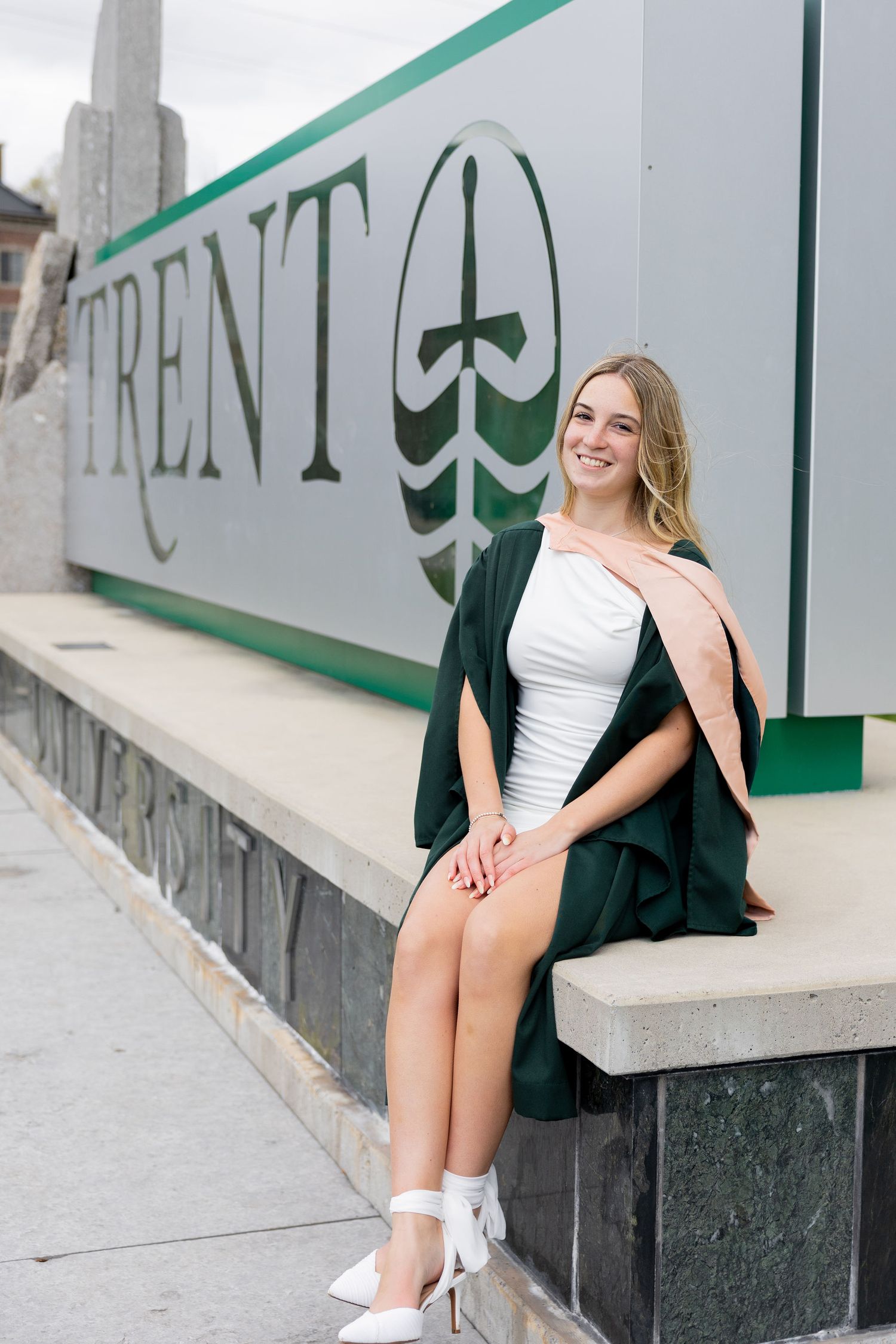 Smiling female graduate in green gown sits beside Trent University sign wearing white dress and heels.
