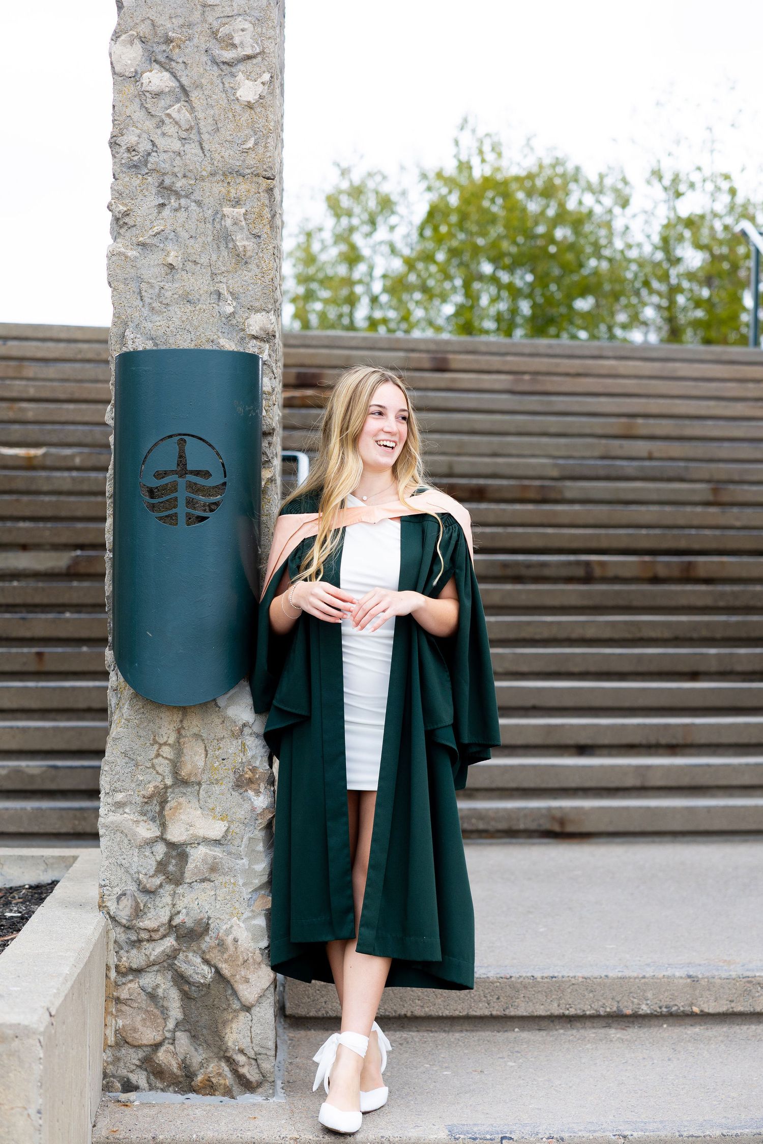 Smiling blonde woman in green graduation gown and white dress poses outdoors near stone pillar and wooden steps.