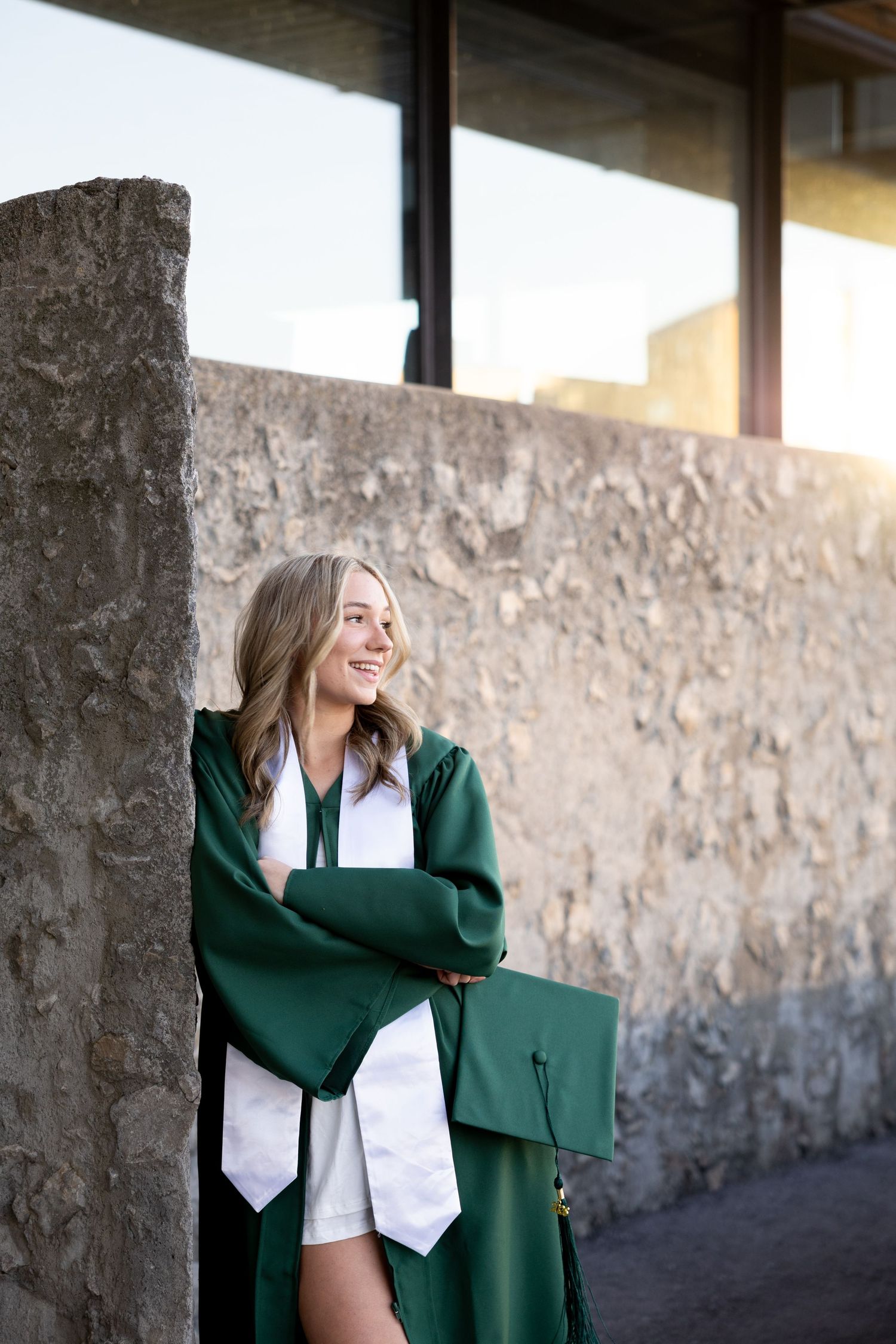 Smiling graduate in green cap and gown leaning against stone wall outdoors, holding diploma with arms crossed.