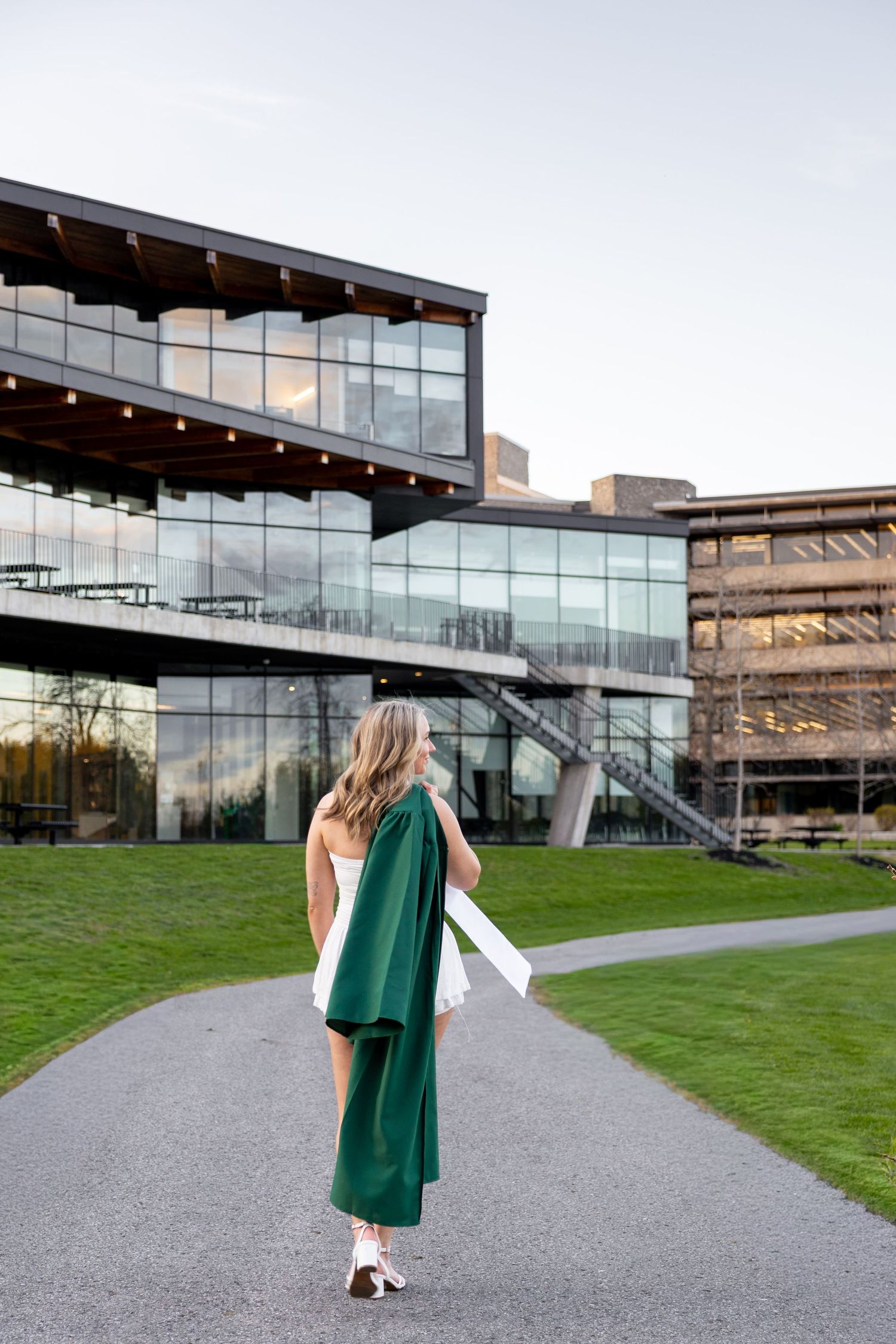A woman in a green graduation gown walks along a path toward a modern glass university building.