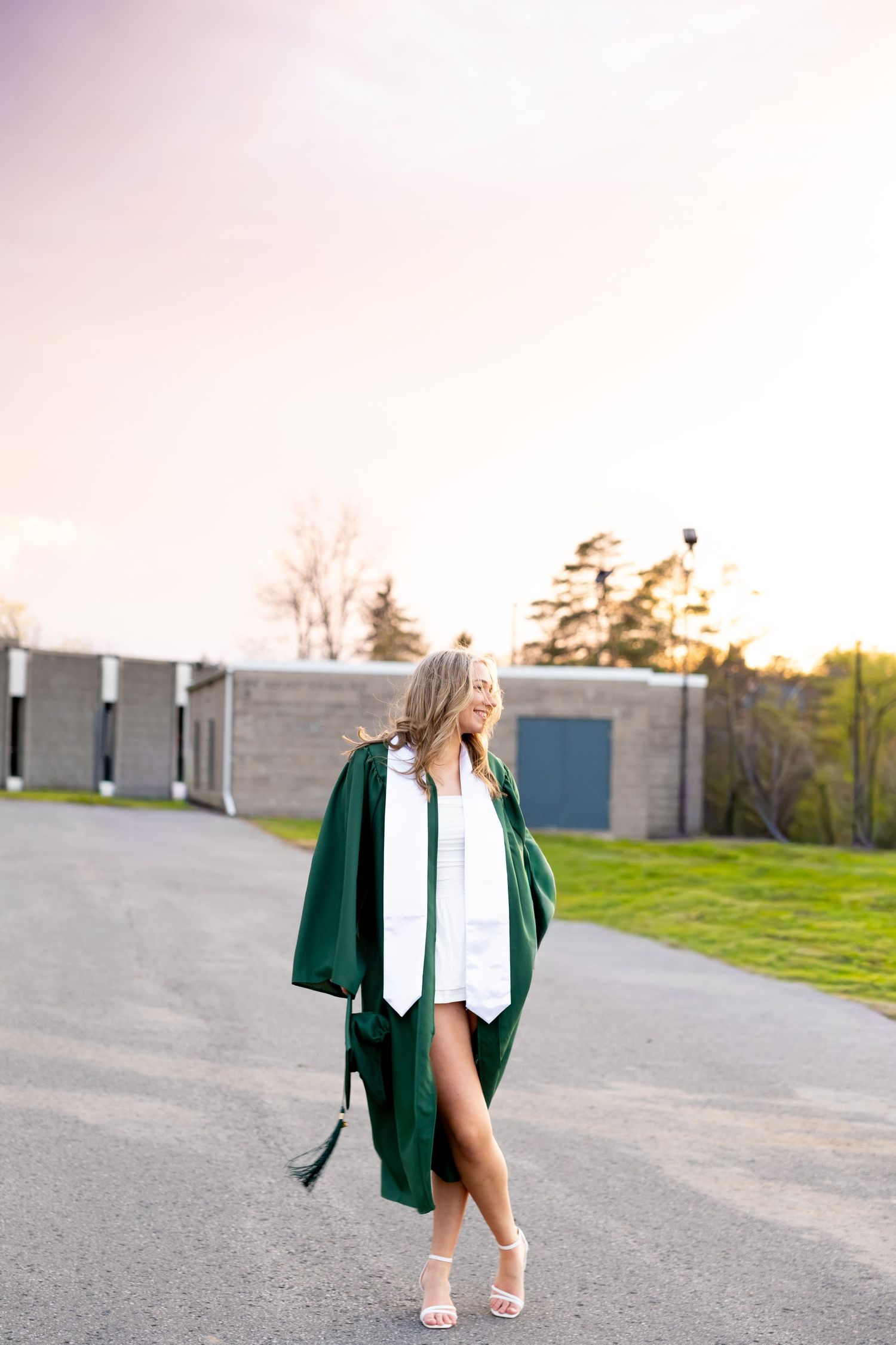 Young woman in green graduation gown and white stole walks on a path, smiling, with a school building in background.