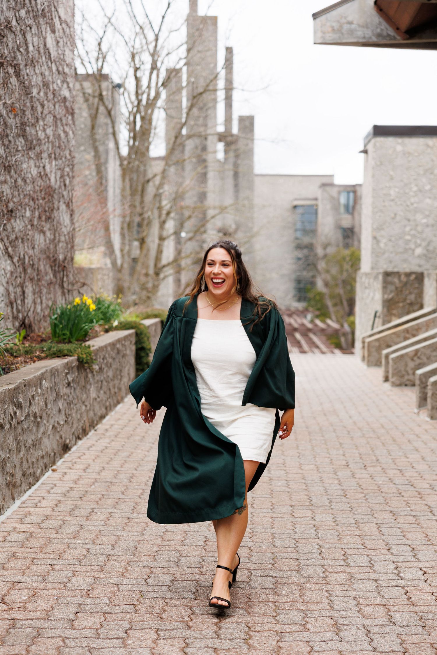 A smiling woman in a white dress and green graduation gown walks along a brick path on a university campus.