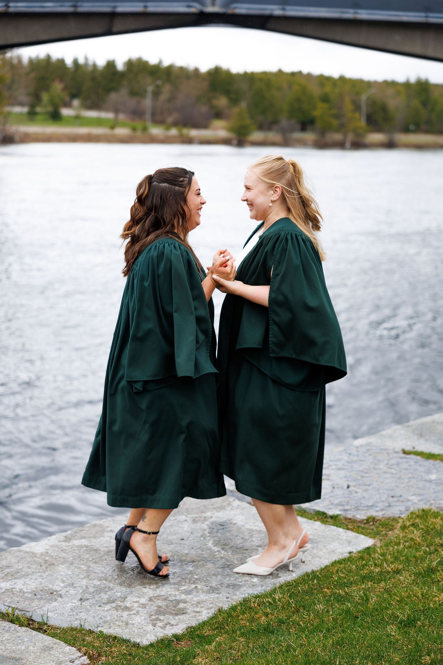 Two women in green graduation gowns laughing together by a river under a bridge.