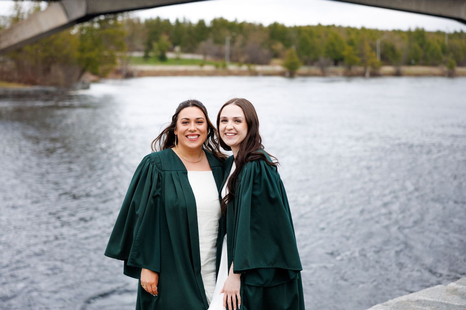 Two graduates in emerald green gowns smile while standing near a scenic waterfront on a sunny day.