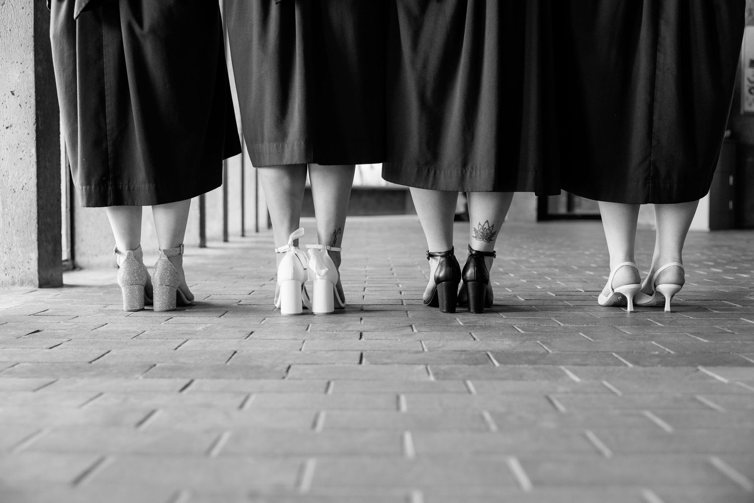 Black and white photo showing a row of women's legs in high heels and skirts standing on a tiled floor.