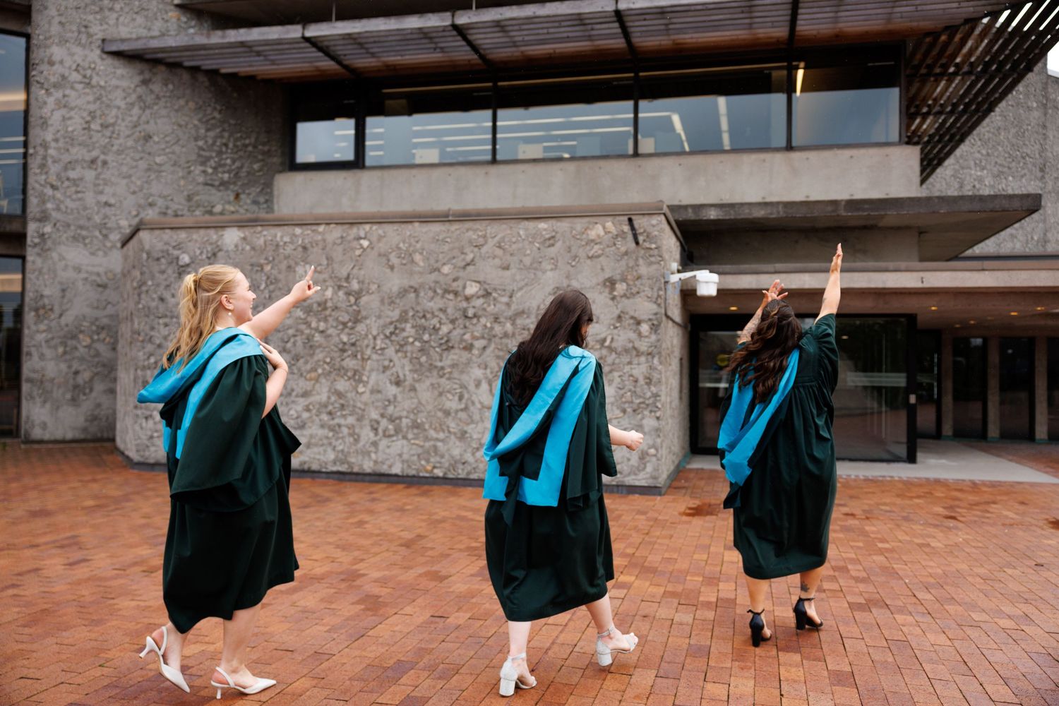 Graduates in dark green robes pose in front of a rustic brick building during commencement celebrations.