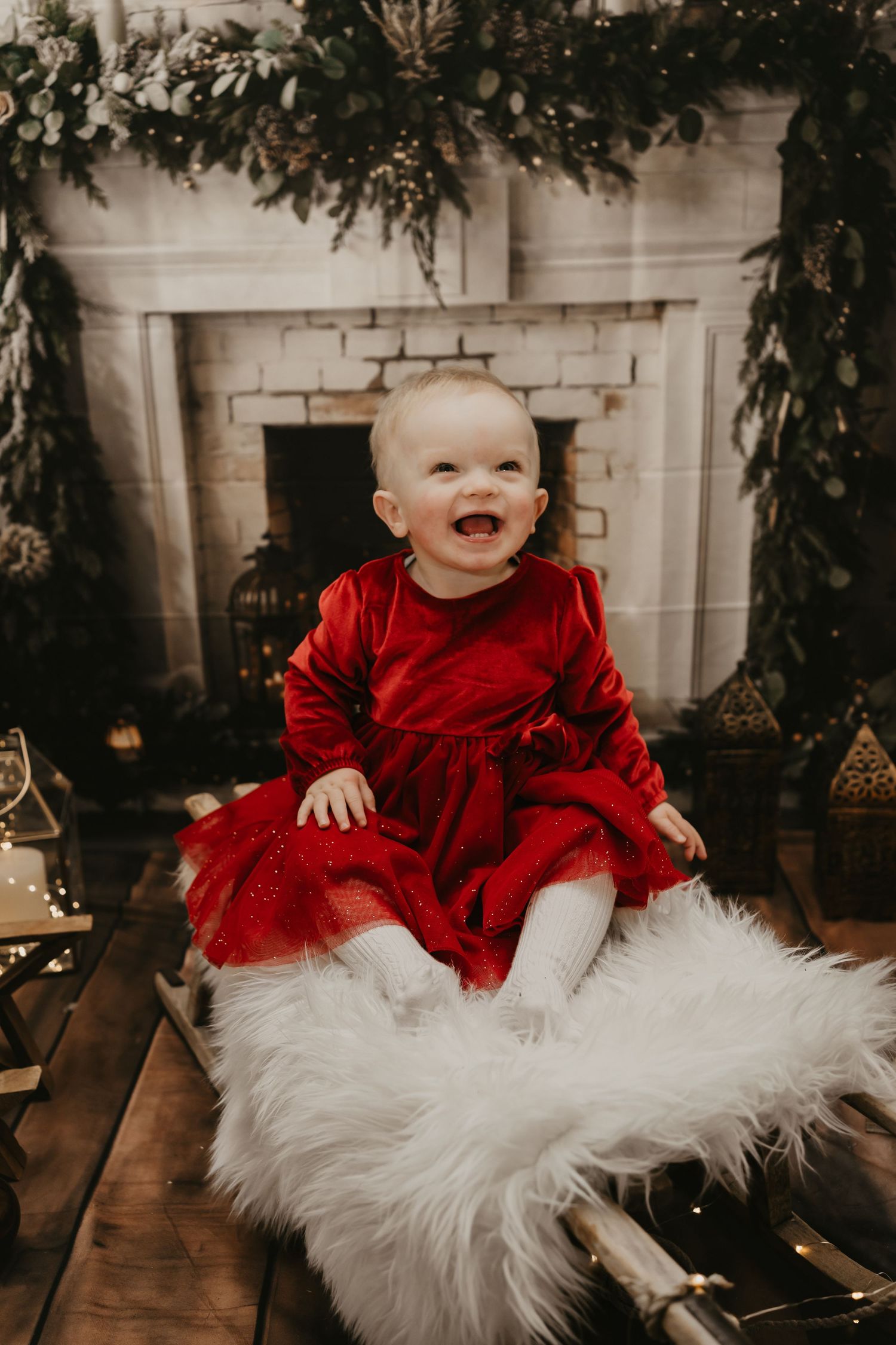 A child in a red dress and white tulle skirt sits in front of a decorated Christmas backdrop with candles and greenery.