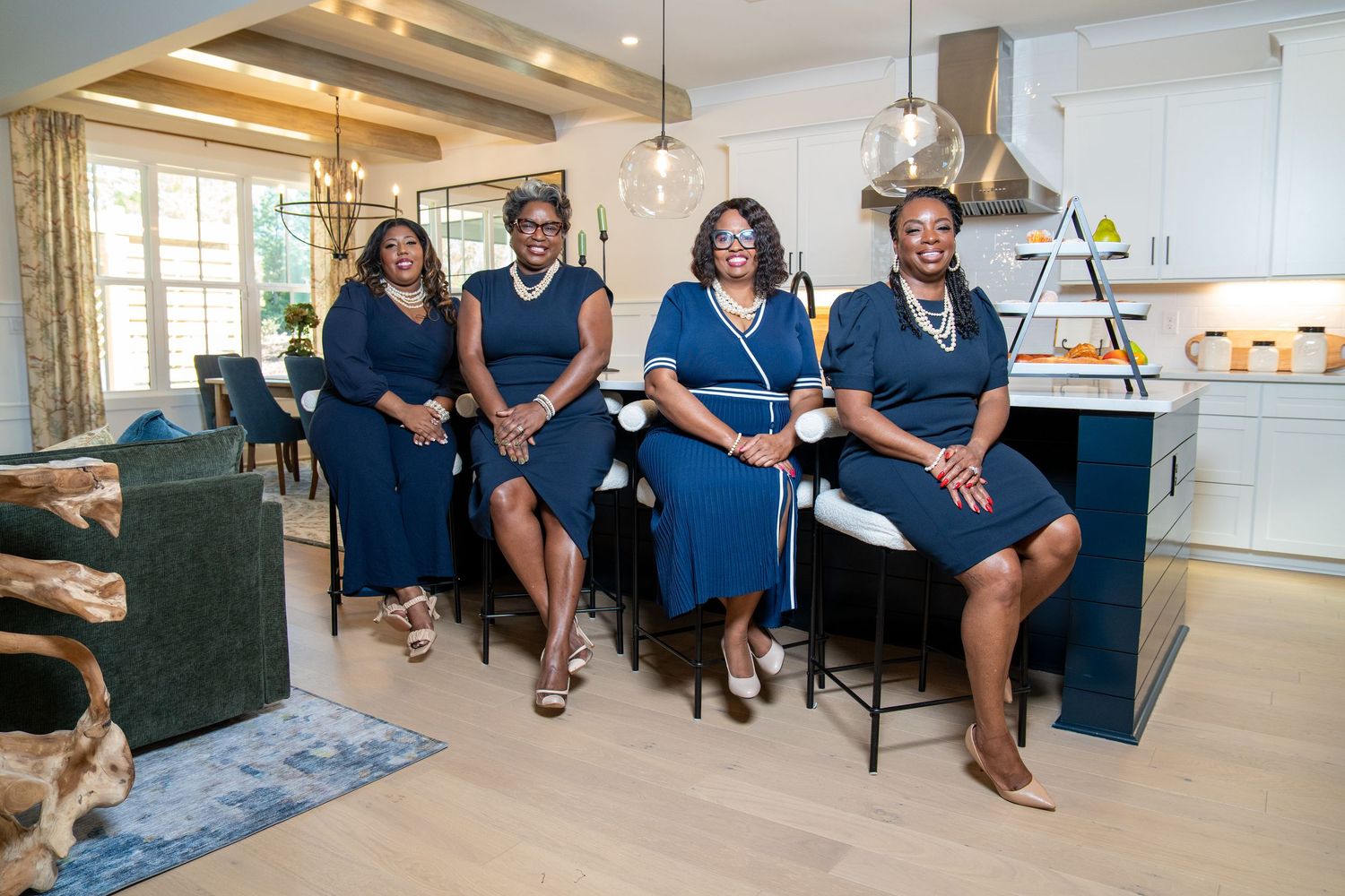 Four women in matching navy blue dresses sit together at a kitchen island in a bright, modern model home.