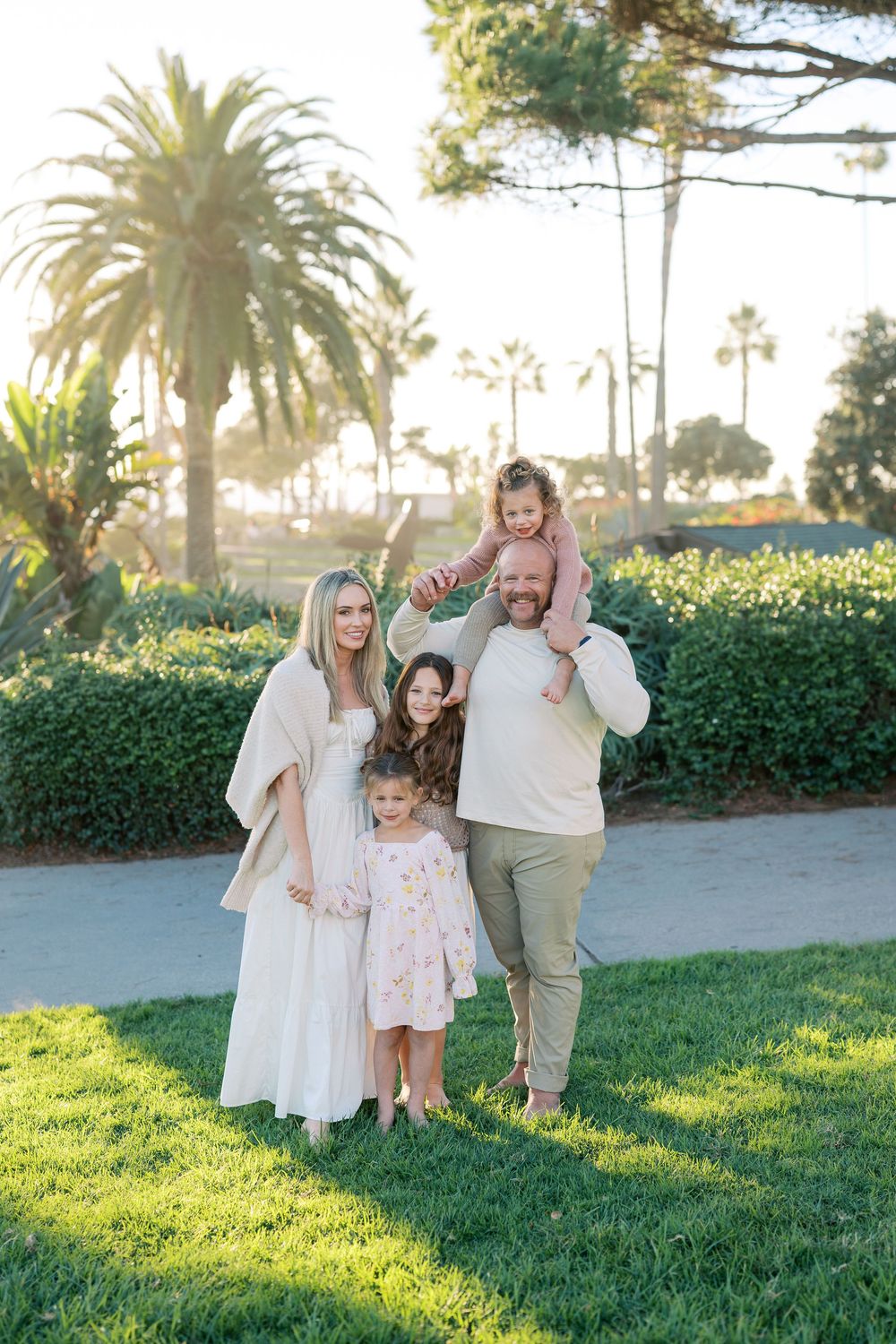 A family in light colored clothing plays together on a grassy lawn with palm trees at sunset.