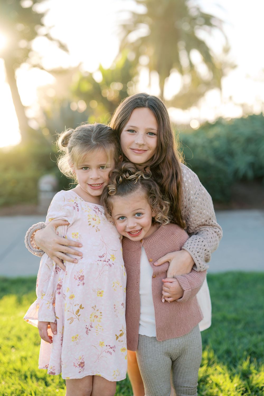 A sweet outdoor family moment captured in warm sunlight on grassy ground wearing soft pink and neutral tones.