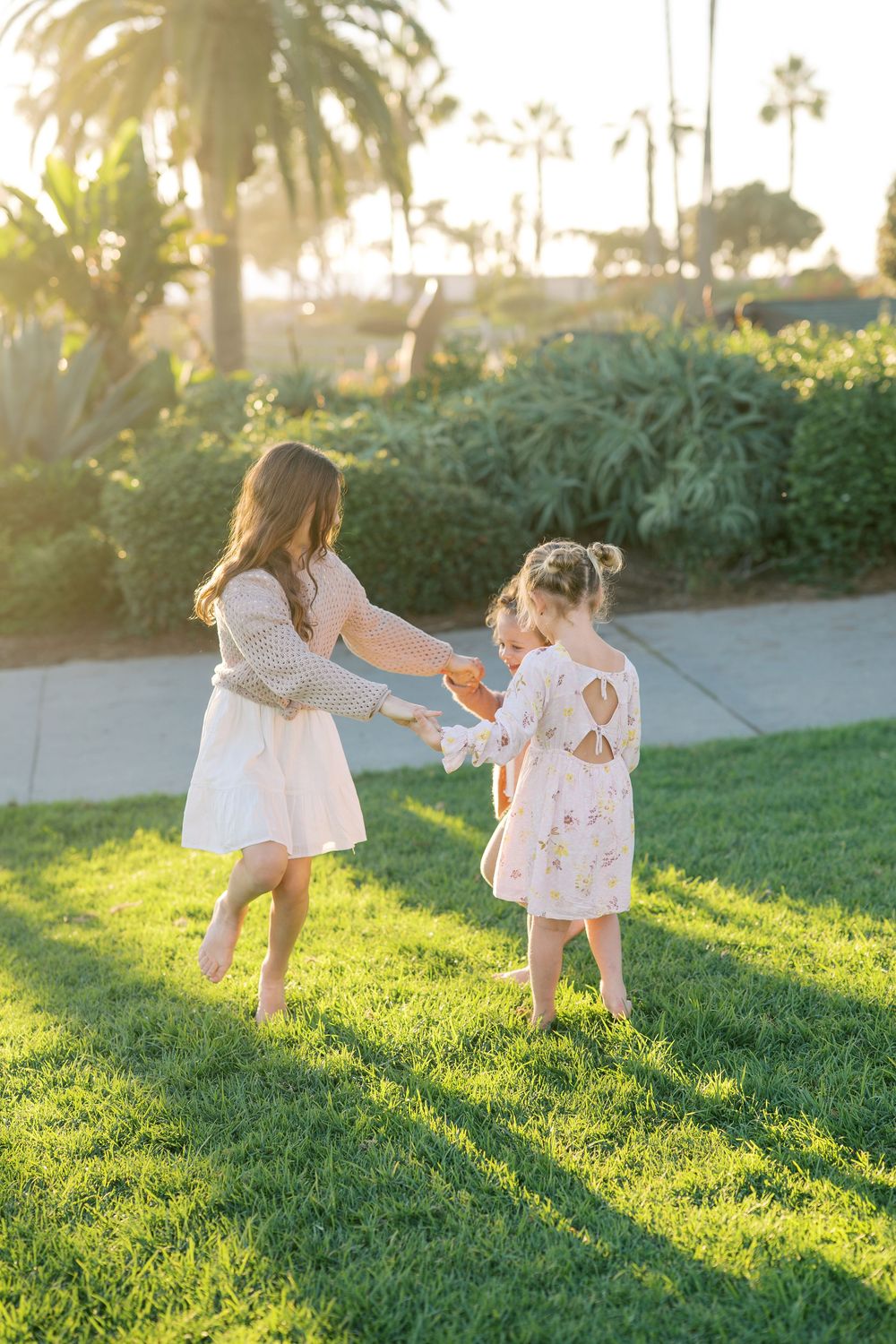 Two children in white dresses play together on a grassy lawn during golden hour with palm trees in the background.