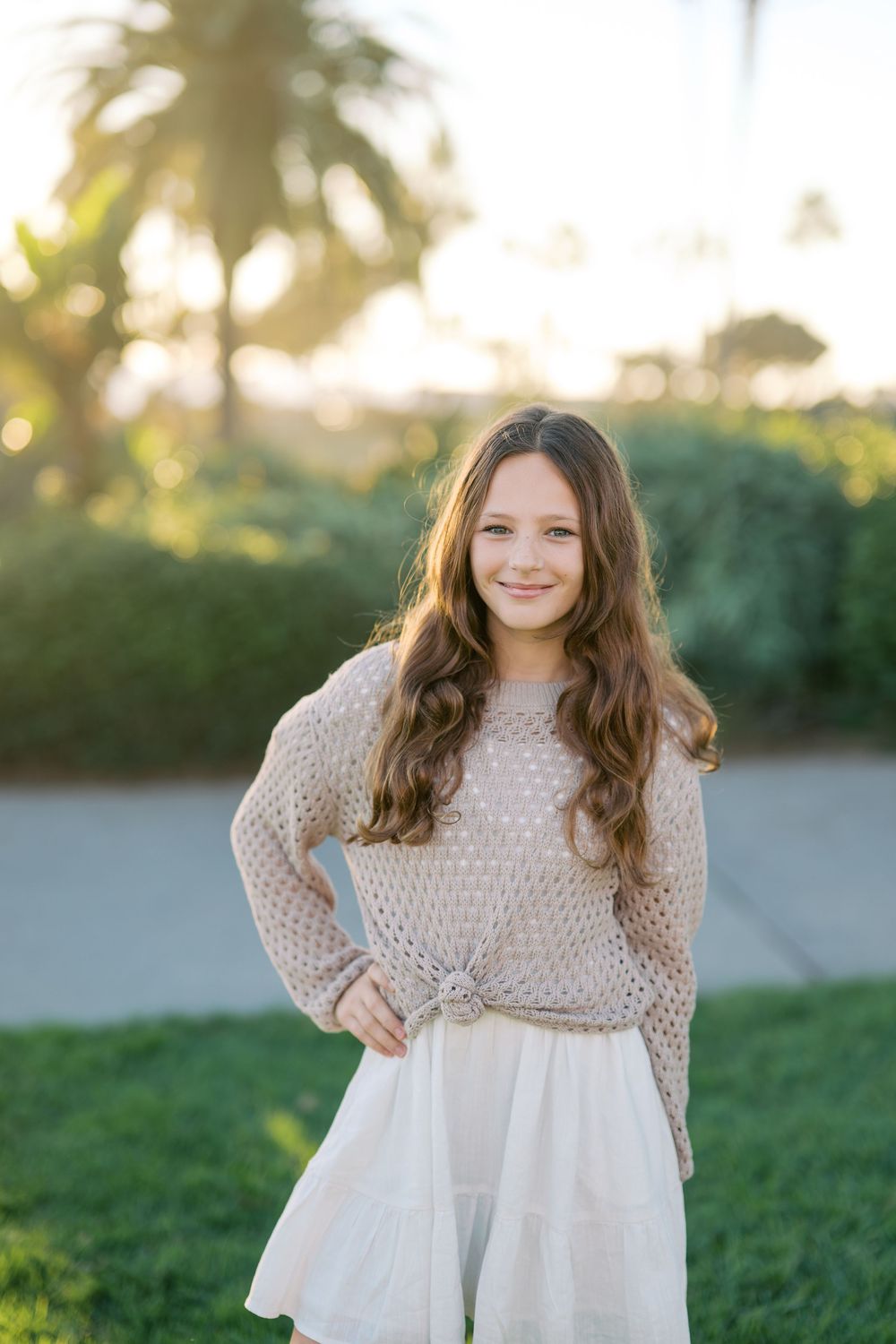 A young girl in a cream knit sweater and white dress twirls in a grassy field during golden hour.