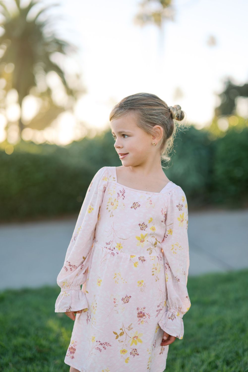 A young child in a pink floral dress stands in a sunlit garden setting.